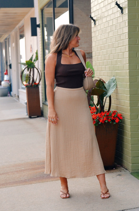 Woman in a black top and beige skirt standing on a sidewalk next to a building with plants.